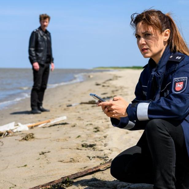 Polizistin kniet am Strand mit Handy, im Hintergrund steht ein Mann am Wasser.