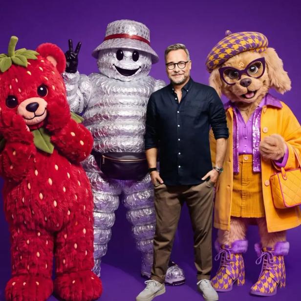 Matthias Opdenhövel steht vor einem violetten Hintergrund zwischen drei Maskottchen aus The Masked Singer.