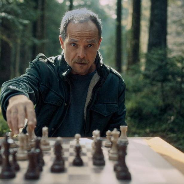 Ein Mann mit Schnurrbart spielt konzentriert Schach an einem Tisch inmitten eines Waldes.