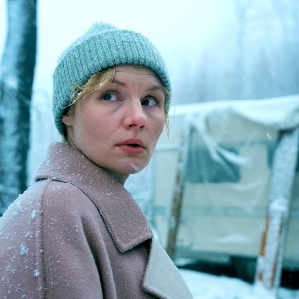 Eine Frau mit hellblauer Mütze und beigem Mantel blickt in einer verschneiten Winterlandschaft vor einem Wohnwagen besorgt zur Seite.