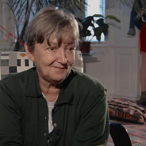 Christine Nöstlinger mit kurzem Haar und dunkelgrünem Pullover sitzt lächelnd in einem Sessel vor einem hellen Raum mit Pflanzen.