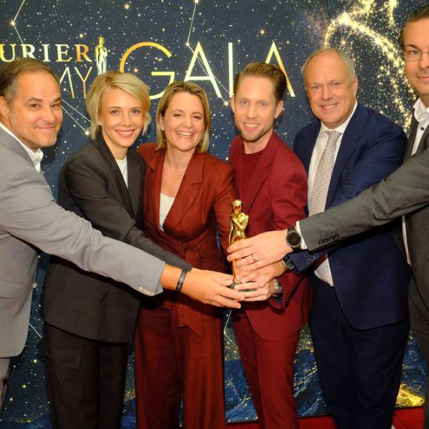 Sechs Personen halten gemeinsam eine goldene Romy-Trophäe vor einer „Kurier Romy Gala“-Wand.