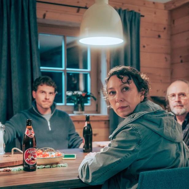 Drei Personen sitzen in einer gemütlichen Holzhütte an einem Tisch mit einer Flasche Alpen-Bier.