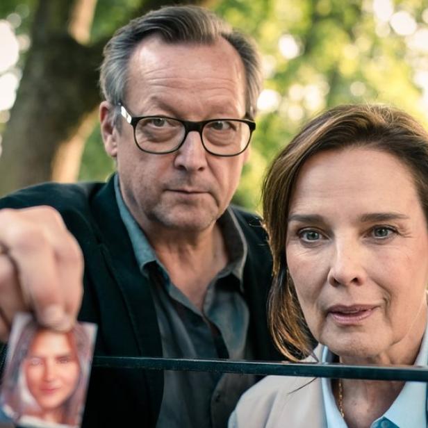 Ein Mann mit Brille und eine Frau schauen ernst in die Kamera, während er ein kleines Porträtfoto einer jungen Frau in die Höhe hält.