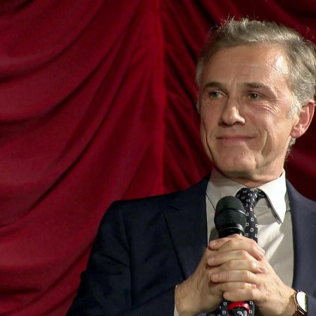 Christoph Waltz spricht in einem Anzug mit Krawatte vor einem roten KIno-Vorhang in ein Mikrofon.