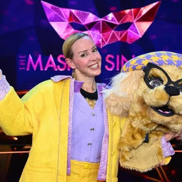 Eine lächelnde Frau posiert neben einer Hundekostüm-Maske auf einer bunten Masked-Singer-Bühne.