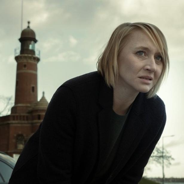 Eine blonde Frau blickt mit besorgtem Ausdruck vor einem Leuchtturm in die Ferne.