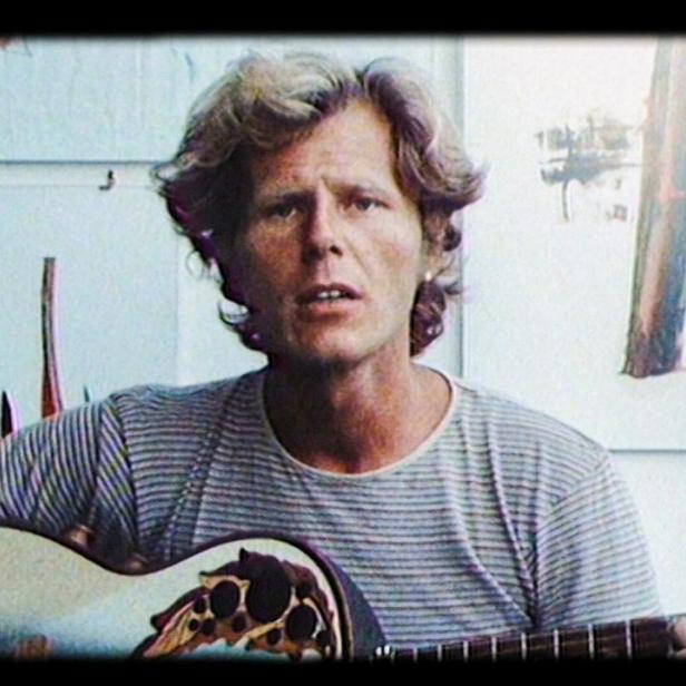 Ein Mann mit lockigem Haar spielt eine Akustikgitarre vor einer weißen Wand mit Zeichnungen.