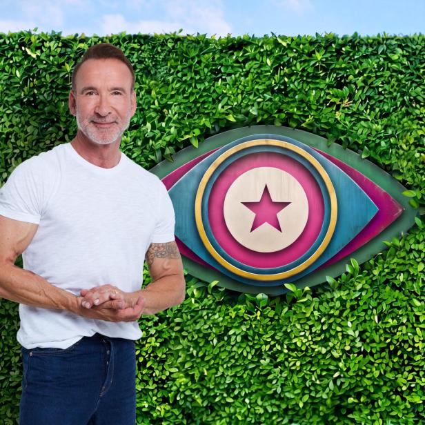 Jochen Bendel steht vor einer grünen Hecke mit dem ikonischen Big-Brother-Auge im Hintergrund.