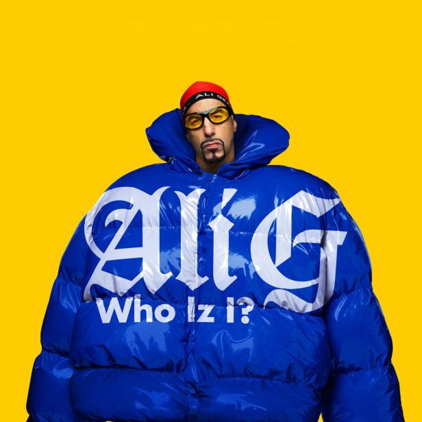 Ali G trägt eine überdimensionale blaue Daunenjacke mit der Aufschrift „Ali G Who Iz I?“ vor einem gelben Hintergrund.