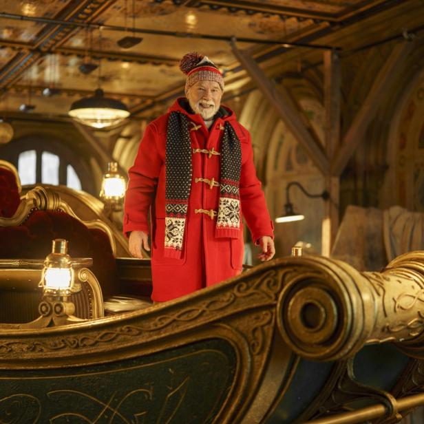 Arnold Schwarzenegger steht als Santa Clause in einem roten Dufflecoat und einer Wintermütze in einem prunkvollen, goldenen Schlitten.