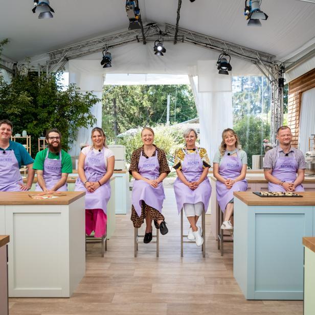 Acht Personen in einer hellen TV-Kochstudio-Küche mit lila Schürzen sitzen und lächeln vor Arbeitsinseln.