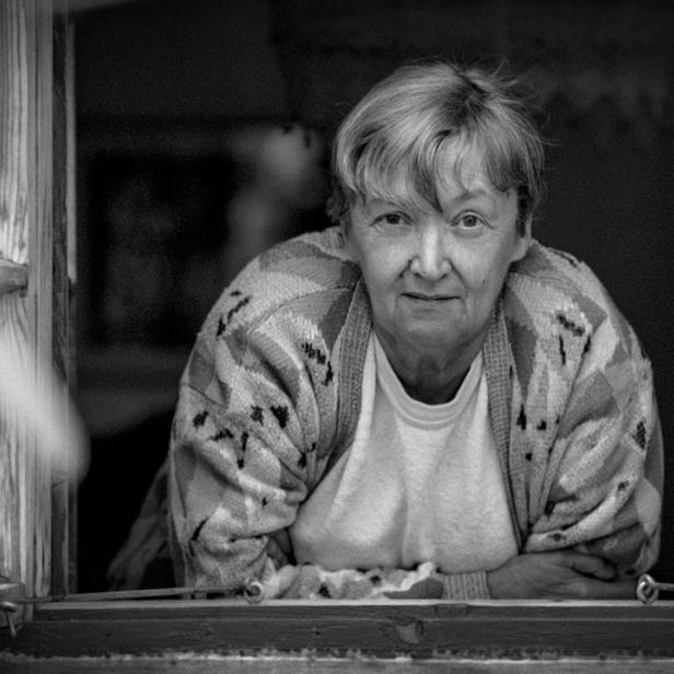 Eine ältere Frau mit kurzem Haar blickt in diesem schwarz-weiß Porträt direkt aus einem geöffneten Fenster nach draußen.