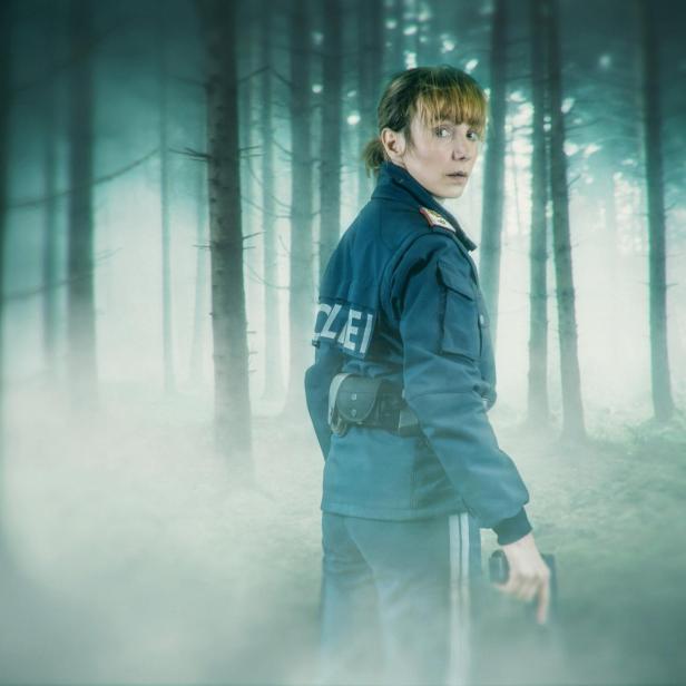 Eine Polizistin in Uniform steht mit einer Pistole in der Hand in einem nebligen Wald und blickt wachsam zurück.