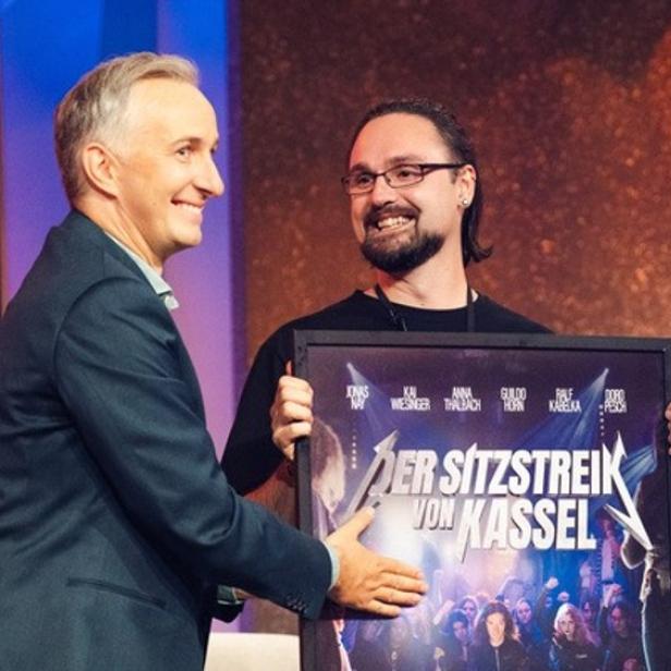 Zwei Männer lächeln, einer überreicht ein gerahmtes Plakat mit dem Titel „Der Sitzstreik von Kassel“.