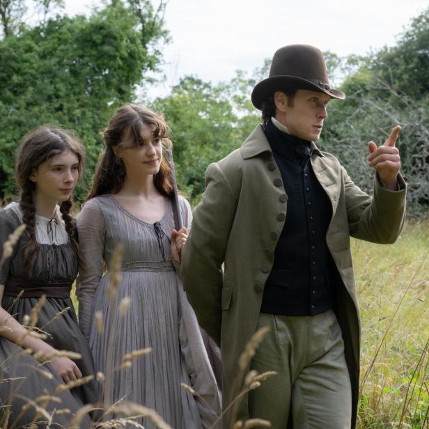 Ein Mann mit Zylinder und zwei Frauen in historischen Kleidern stehen in einer Wiese, während der Mann nach vorne zeigt.