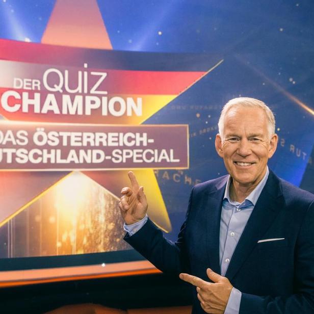 Johannes B. Kerner präsentiert vor einer Studiowand mit dem Logo von „Der Quiz-Champion – Das Österreich-Deutschland-Special“.