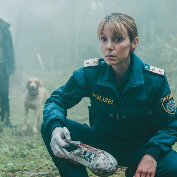Eine Polizistin in dunkler Uniform hält im Wald einen blutigen Schuh, während im Hintergrund ein Mann mit einem Hund steht.
