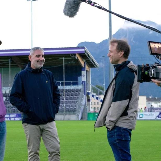 Ein Kamerateam filmt drei Männer im Gespräch auf einem Fußballplatz vor einer Tribüne.