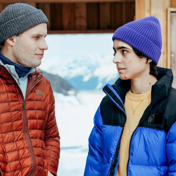 Ein Mann mit grauer Mütze und oranger Jacke blickt einen Jüngeren mit blauer Mütze und farbiger Jacke an, der leichte Verletzungen im Gesicht hat.