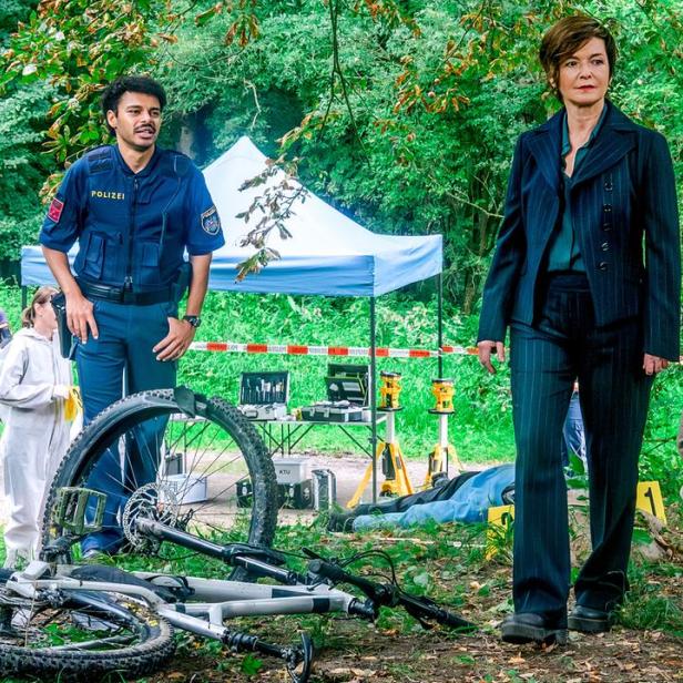 Einsatzkräfte und Polizisten untersuchen im Wald einen Tatort mit einem liegenden Fahrrad und abgesperrtem Bereich.