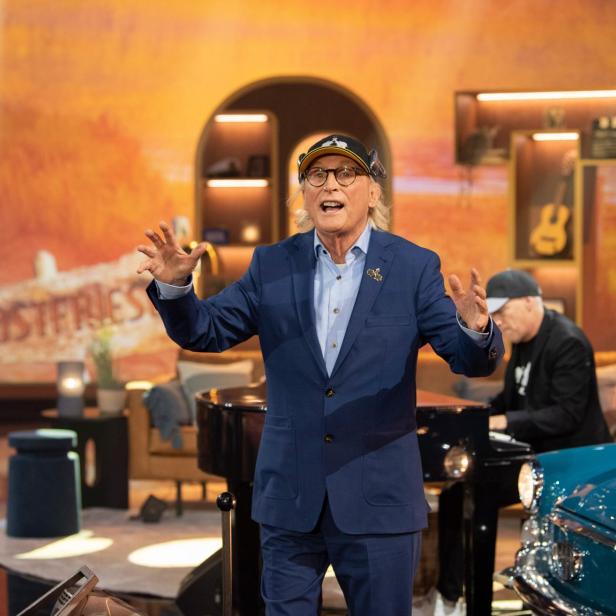 Otto Waalkes im blauen Anzug und mit Kappe spricht gestikulierend in einem TV-Studio, daneben sitzt ein Pianist am Flügel.