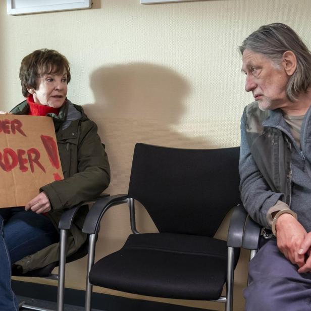 Zwei ältere Personen sitzen in einem Warteraum; die Frau hält ein Schild mit der Aufschrift „Einmal Mörder, immer Mörder“.