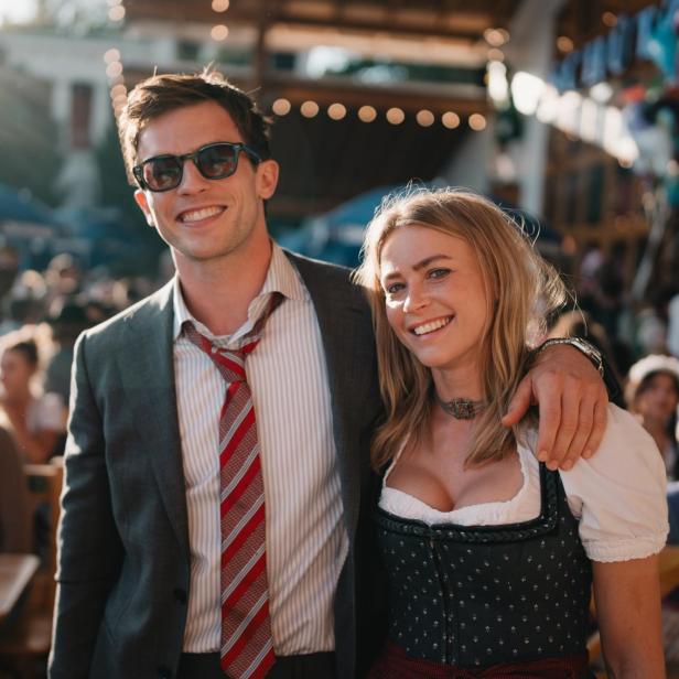 Ein lächelndes Paar steht Arm in Arm auf einem Fest; er trägt Anzug und Krawatte, sie ein traditionelles Dirndl.