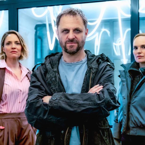 Ein Mann mit verschränkten Armen steht im Vordergrund, flankiert von zwei Frauen vor einer Glaswand mit hellen Neonlichtern.